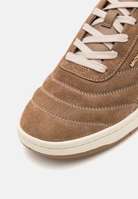 Gros plan d'une basket en daim marron avec des lacets crème, des surpiqûres blanches et une semelle en caoutchouc blanche sur un fond clair.
