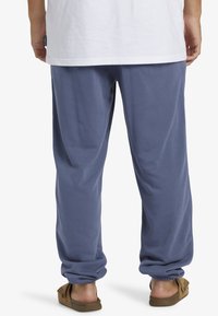 Blå bomullssweatpants med elastiska mudd, avslappnad passform, svag glans, kombinerat med bruna slip-on sandaler, bakvy, vit t-shirt synlig.