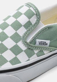 Slinkskor utan snörning med en grön och vit rutig ovandel i canvas. Har en vit gummisula och Vans-logotyp. Inskränkt sömnadsdetalj.