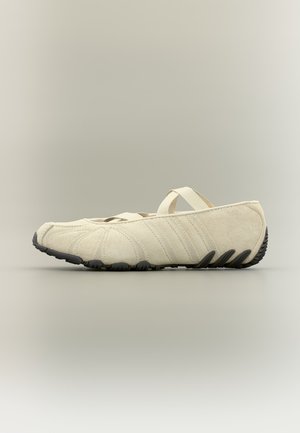Beige blødt stof slip-on sko med tynd elastisk rem og sort tekstureret sål, vist fra siden på en neutral baggrund.