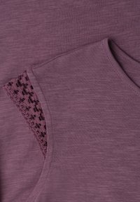 Manche de chemise en tissu violet avec détail en dentelle au crochet près de l'épaule, montrant une couture fine et un motif tricoté texturé.