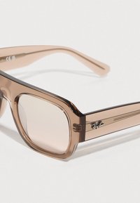 Καφέ αποχρώσεις γυαλιών ηλίου με τετράγωνα φακούς, διάφανο σκελετό από οξικό οξύ και μεταλλικές λεπτομέρειες. Διαθέτουν διακριτικό λογότυπο Ray-Ban στο μπράτσο.