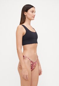 Crni bikini top s širokim trakama; u kombinaciji s donjim dijelom bikinija s cvjetnim uzorkom u nijansama ružičaste i crne, koji ima podesive vezice i tanke bočne trake.