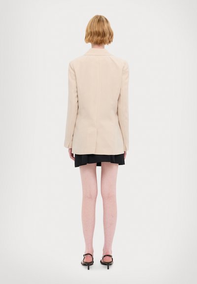 Blazer beige ajusté avec des épaules structurées et une fente arrière unique, associé à une jupe plissée noire et des escarpins à lanières.