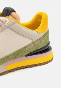 Baskets de sport avec tissu beige, accent en daim vert et semelle en caoutchouc jaune, rose et noire. Comprend un logo et une doublure texturée.