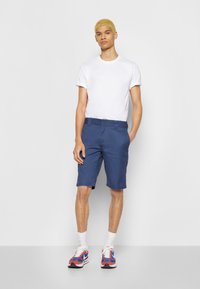 Calções de algodão azuis com corte direto, apresentando bolsos laterais e passadores para cinto, combinados com uma t-shirt branca de decote redondo e sapatilhas coloridas.