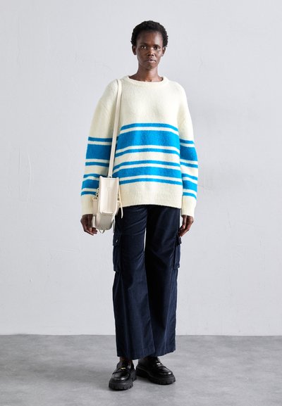 Pulover oversized de culoare crem, cu dungi orizontale albastre și albe, purtat cu pantaloni cargo bleumarin și pantofi cu toc negru. O geantă mică de culoare crem este purtată încrucișat.