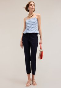 Top peplum a righe blu chiaro con spalline sottili, abbinato a pantaloni navy sartoriali. La modella tiene una borsa multicolore e indossa tacchi bianchi a blocco.