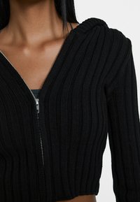 Svart ribbad zip-up huvtröja med en figursydd design, texturerat stickat tyg och rundad huva. Har en silverfärgad dragkedja. Kort cropped längd.