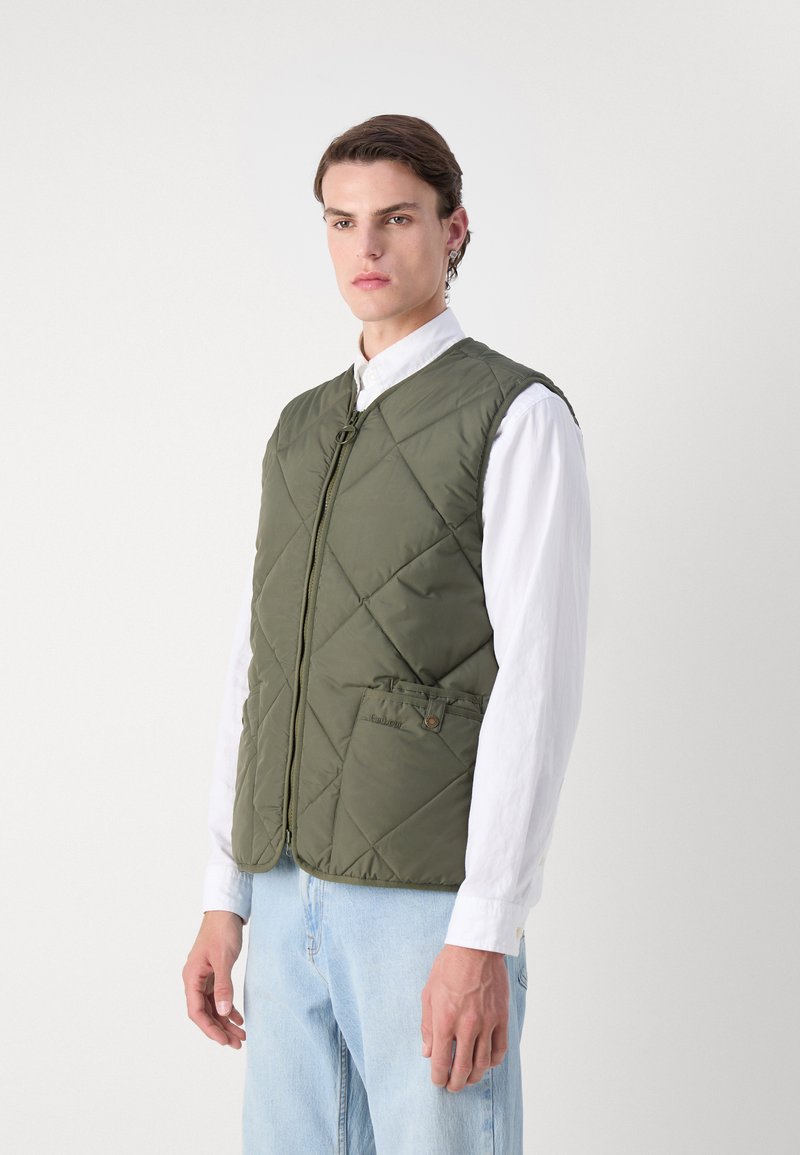 Gestepptes olivgrünes Weste mit einem Frontreißverschluss, seitlichen Taschen und einer leichten Textur, getragen über einem weißen Kragenhemd und hellblauen Jeans.