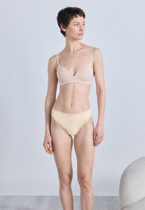 Femme aux cheveux bruns courts portant un soutien-gorge couleur chair et une culotte beige bordée de dentelle, debout devant un fond clair uni.