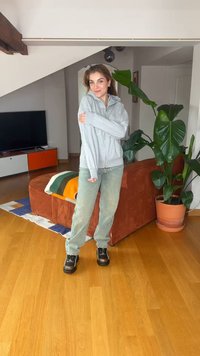 Νεαρή γυναίκα με ανοιχτό γκρι φούτερ και πράσινο παντελόνι στέκεται χαμογελαστή στο σαλόνι με πορτοκαλί καναπέ, μεγάλο φυτό και κονσόλα τηλεόρασης.