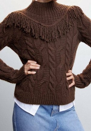Brun kabelstrikket sweater med frynse-detalje på brystet, båret over en hvid skjorte og kombineret med lyseblå jeans, hænder hvilende på hofterne.