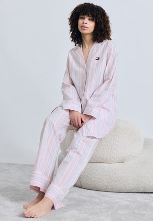 Frau mit lockigem Haar sitzt barfuß auf einem strukturierten cremefarbenen Pouf und trägt einen rosa-weiß gestreiften Pyjama mit langen Ärmeln und kleinem Logo.