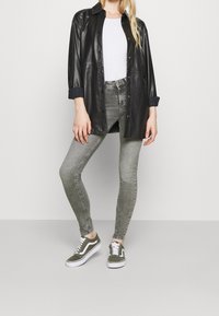 Zwarte faux leren knop-overhemd, witte fitted t-shirt, grijze skinny jeans en groene Vans sneakers met witte accenten. Gladde textuur, casual stijl.