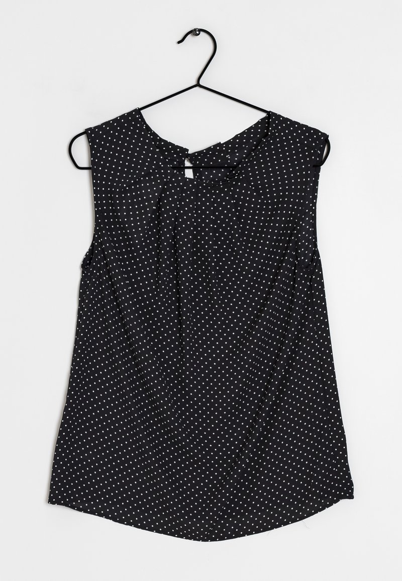 Blouse noire sans manches à pois blancs accrochée sur un cintre noir sur un fond blanc.