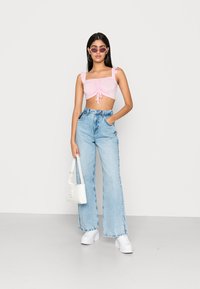 Top cropped rosa com alças em babados, detalhe de laço na frente, combinado com jeans de perna larga azul claro e ténis brancos. Uma bolsa branca completa o visual.