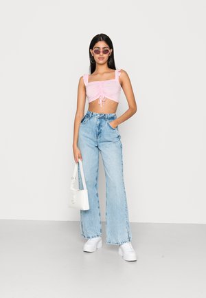 Top cropped rosa com alças em babados, detalhe de laço na frente, combinado com jeans de perna larga azul claro e ténis brancos. Uma bolsa branca completa o visual.