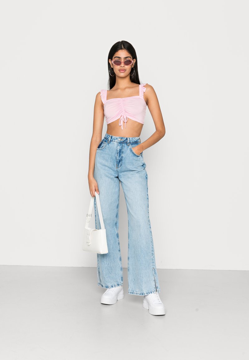 Top cropped rosa com alças em babados, detalhe de laço na frente, combinado com jeans de perna larga azul claro e ténis brancos. Uma bolsa branca completa o visual.
