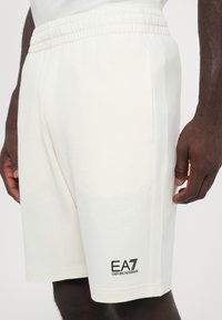 Svijetlosmeđi šorts s elastičnom strukom, bočnim džepovima i crnim EA7 Emporio Armani logotipom blizu rubova. Glatka tekstura tkanine.
