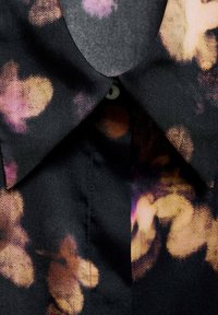 Blouse noire à motif floral dans des tons de violet et d'orange, avec un col pointu et une fermeture à boutons au niveau du cou.