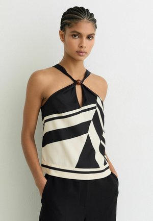 Kobieta z zaplecionymi włosami, ubrana w czarno-biały geometryczny top na szyję z drewnianym pierścieniem, ręce w kieszeniach czarnych spodni, neutralne tło.