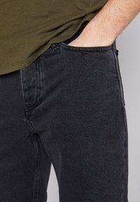 Melnas džinsu bikses ar šauru piegriezumu, kurām ir klasiskā piecu kabatu dizains un smalki izšuvumi. Apvienots ar zaļu t-kreklu.