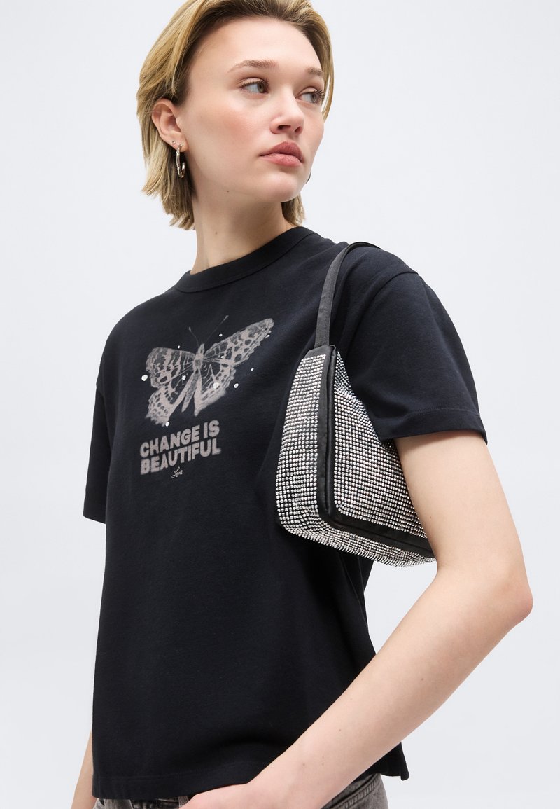 Jeune personne portant un t-shirt noir avec un papillon et le texte "LE CHANGEMENT EST BEAUTÉ", portant un sac à épaule orné de strass, regardant sur le côté.