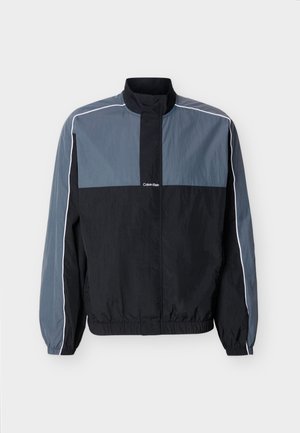 Veste légère zippée pour homme en noir et gris avec col montant, poignets et ourlet élastiques, et passepoil blanc le long des manches et des épaules.