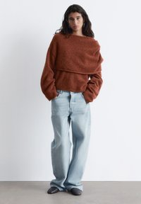 Rust-colored, soft sweater with a layered front and wide sleeves, paired with light blue high-waisted denim jeans, and gray slip-on shoes.