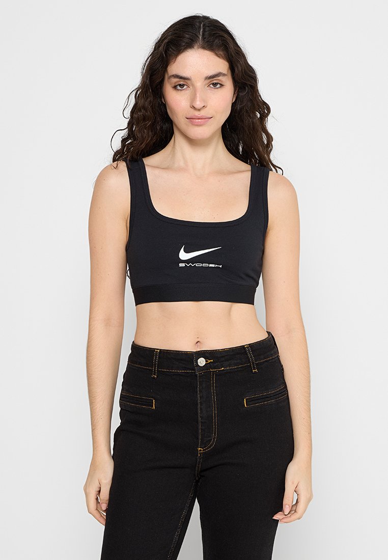 Soutien-gorge de sport noir avec un décolleté carré, portant un logo Nike blanc et le texte "Swoosh". Associé à un jean noir taille haute.