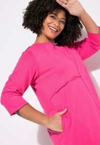 Abito di cotone rosa con maniche a tre quarti, caratterizzato da un collo rotondo, tasca frontale e dettagli a coste lungo le cuciture.