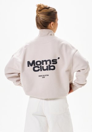 Kobieta z włosami związanymi w kok, ubrana w jasnobeżową bluzę z napisem „Moms' club Mirons Collection 2026” na plecach, w białych spodniach.