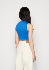 Kvinna som bär en ärmlös ljusblå crop top och högmidjade vida vita jeans, stående med ryggen mot kameran.