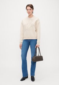 Μπεζ πουλόβερ με κέντημα ροζ, μπλε καμπάνα τζιν, μαύρες μποτάκια έως τον αστράγαλο και σκούρο καφέ τσάντα με διπλές λαβές.