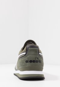 Zapatilla verde oliva hecha de ante con acentos en blanco y negro; cuenta con un cuello acolchado, suela texturizada y un logotipo en el talón.