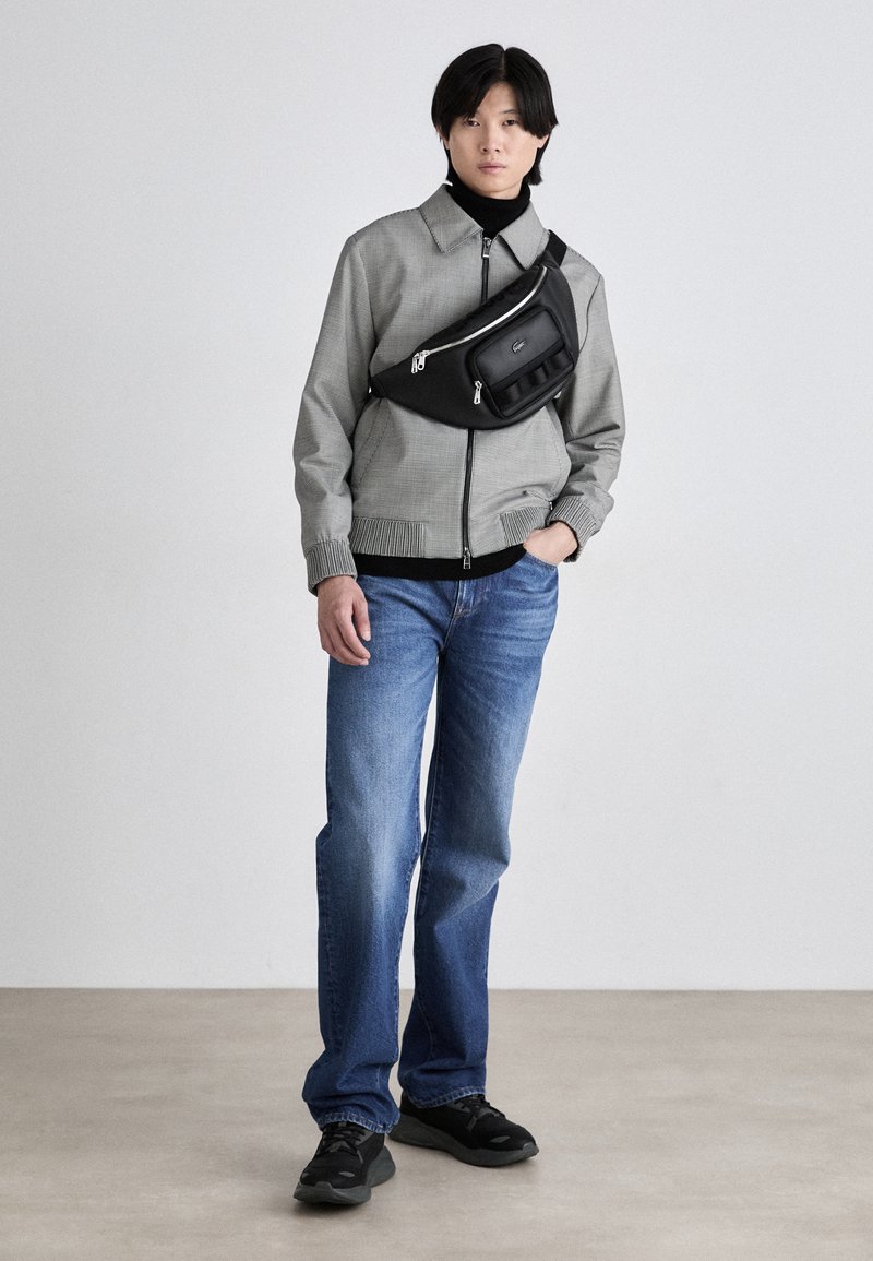 Veste bomber à motifs gris, sac banane noir, col roulé noir, jean bleu et baskets noires. Le mannequin se tient devant un fond neutre.