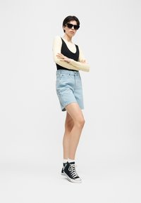 Άνδρας που φοράει μαύρο αμάνικο τοπ πάνω από μπεζ πουκάμισο με μακριά μανίκια, ανοιχτόχρωμα τζιν σορτς, μαύρα ψηλά αθλητικά παπούτσια και μαύρα γυαλιά ηλίου, στέκεται με τα χέρια σταυρωμένα.