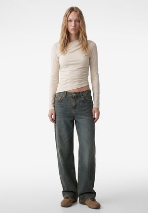 Mujer joven con el pelo largo y rubio, vistiendo una camiseta ajustada de manga larga beige, vaqueros grises desgastados y sueltos, y zapatos marrones, de pie sobre un fondo blanco.