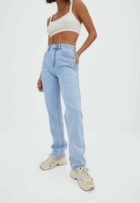 Femme portant un jean taille haute bleu clair, un crop top crème et des baskets beige, se tenant devant un fond blanc uni.