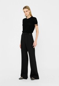 Schwarzes Strickoberteil mit kurzen Ärmeln und Rautenmuster, kombiniert mit hoch taillierten schwarzen Schlaghosen und grauen flachen Schuhen.