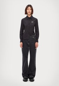 Camicia nera con bottoni e colletto, con un logo sul lato sinistro del petto. Abbinata a jeans in denim scuri a gamba larga e scarpe nere massicce.
