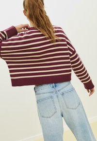 Personne aux cheveux longs portant un pull à rayures bordeaux et beige et un jean taille haute bleu clair, vue de dos avec un bras levé.