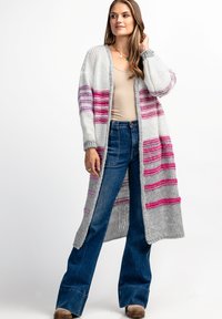 Długi szary sweter z dzianiny z różowymi i fioletowymi paskami, z otwartym przodem i ściągaczami przy rękawach, w połączeniu z jasnoniebieskimi dżinsami dzwonami.
