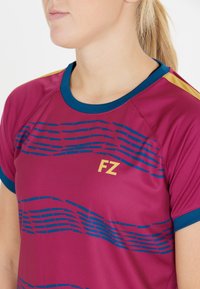 Nainen, joka on pukeutunut marooninväriseen urheilupaitaan, jossa on sinisiä aaltokuvioita, siniset kaulukset ja hihansuut sekä keltainen "FZ"-logo rinnassa.