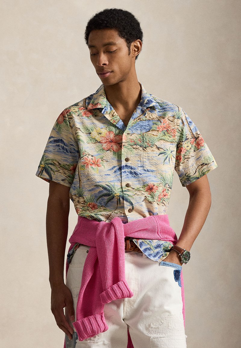 Muž nosi kvetovanú krátkosrúbovú košeľu na gombíky, biele nohavice s náplasťami, ružový sveter zviazaný okolo pása a náramkové hodinky.