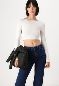 Top corto de manga larga blanco combinado con jeans de tiro alto azul oscuro. La modelo sostiene una chaqueta acolchada negra. Fondo simple.