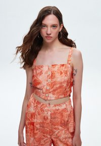 Tweekleding bestaande uit een cropped, oranje top met een bloemenpatroon en knoopsluiting, gecombineerd met bijpassende high-waisted broek.