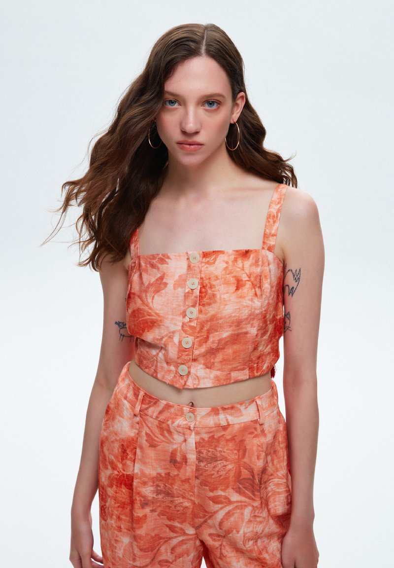 Tweekleding bestaande uit een cropped, oranje top met een bloemenpatroon en knoopsluiting, gecombineerd met bijpassende high-waisted broek.