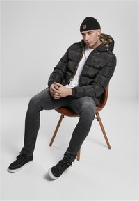 Homme portant un bonnet noir, une doudoune noire avec une capuche camouflage, une chemise blanche, un jean gris et des baskets noires, assis sur une chaise marron.